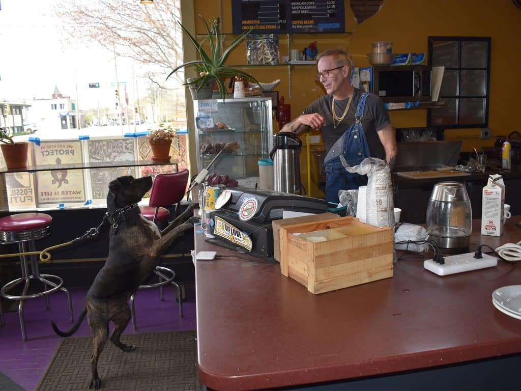 Photo of Patrick Downey, right, behind the counter, looking at a medium-sized dog with it's paws on the other side of the counter, waiting expectantly. 