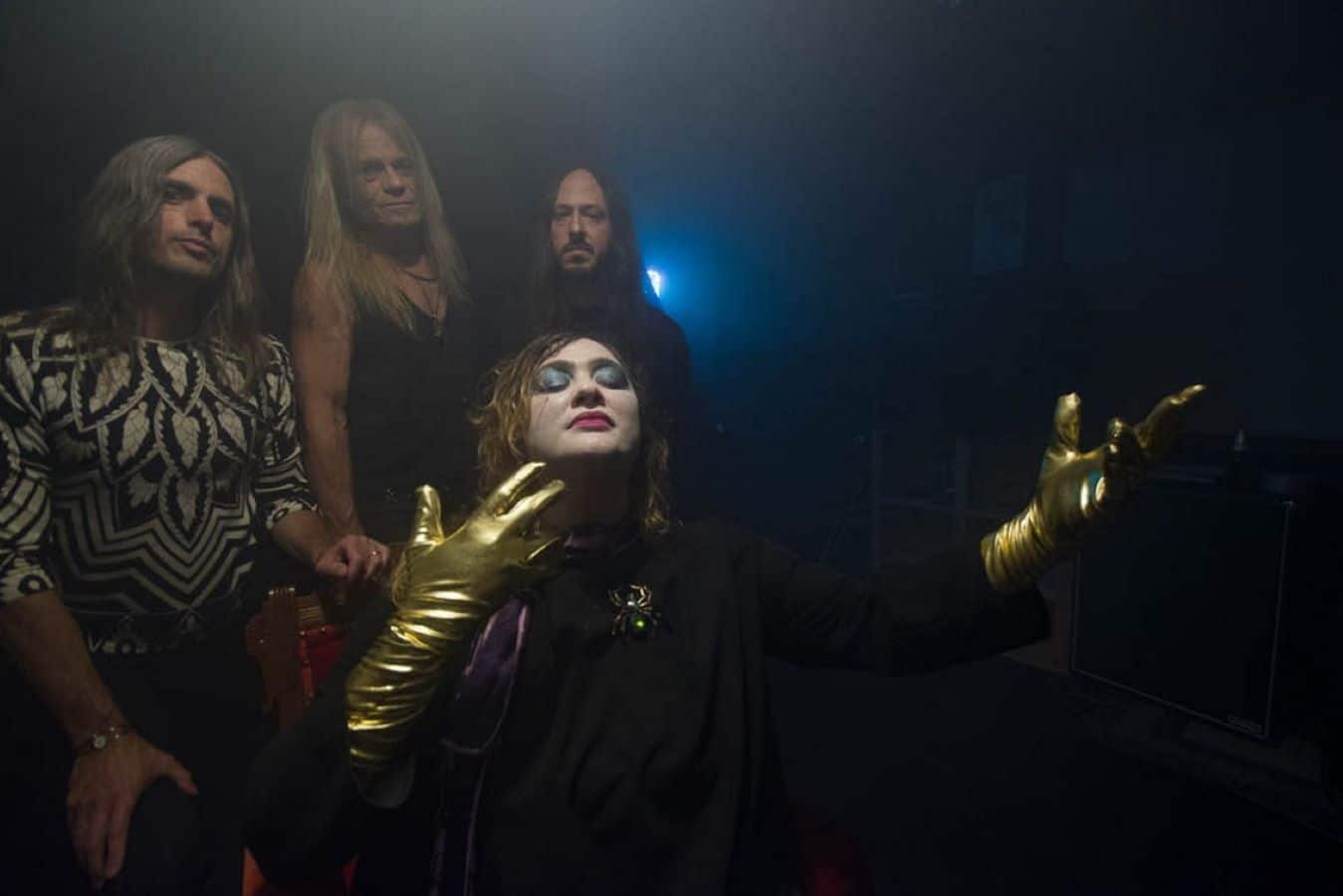 Three members of the band Vanishing Kids stand shoulder to shoulder in the background. From Left to Right: Jason Hartman, Jerry Sofran, and Nick Johnson. In the foreground, Nikki Drohomyreky kneels, with her eyes closed. She's wearing gold gloves and his her arms stretched out dramatically. Behind the band, who are dimly lit, there is a soft blue light visible in the center of the image, partially shielded by Johnson's shoulder.