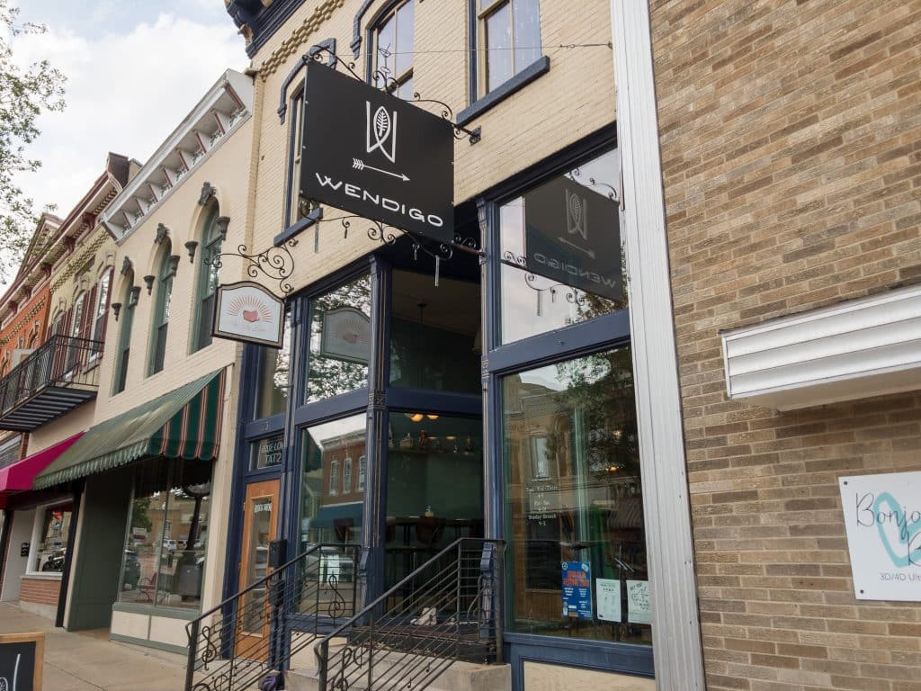 A narrow shopfront with a small concrete staircase leads up towards a black sign that reads "Wendigo" in white capital letters. An arrow pointing towards the entrance and a fish-like sign with a tree at the center appear above the lettering.