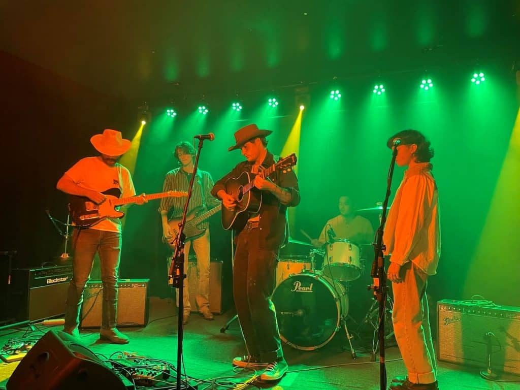 Blue County pistol is shown performing live on a decently large stage. Green lights cascade down on the band. From Left to Right: Gunnar Smith, Vincent Dunn, Jasper Nelson, Luis Acosta Jr., and Mac Saunders. Smith and Nelson are both wearing cowboy hats.