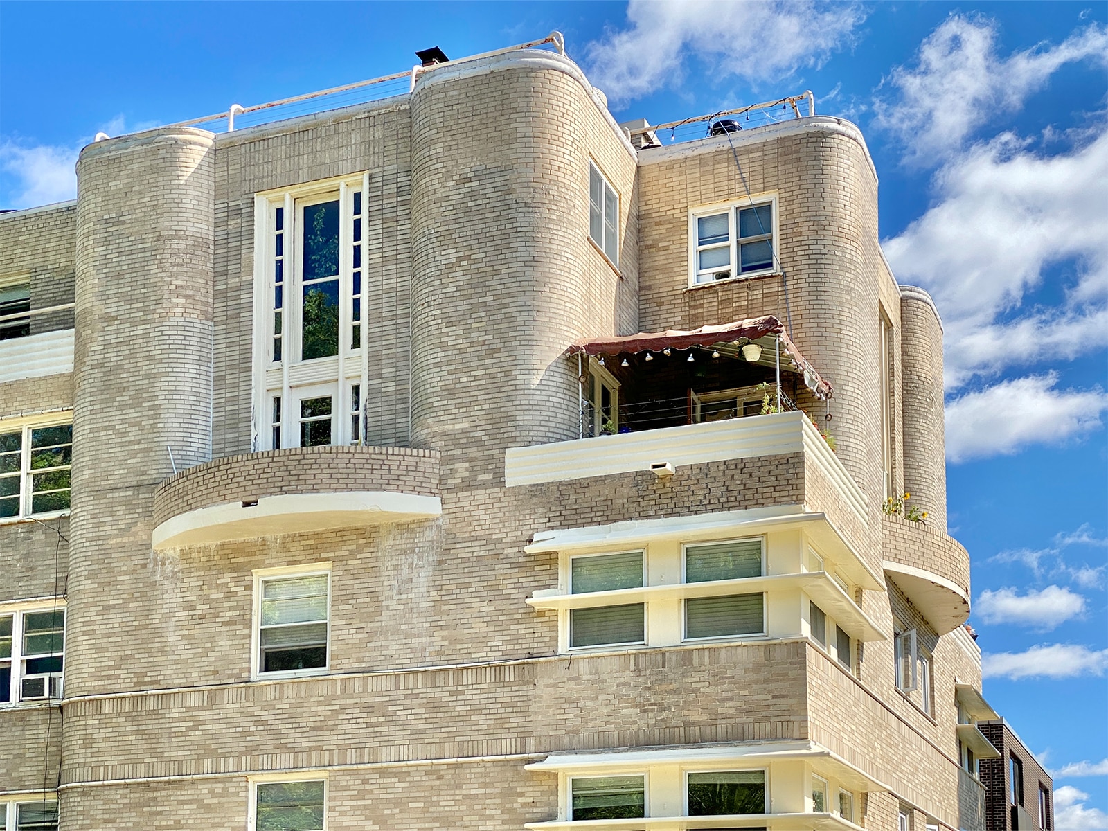 A photo shows the Quisling Terrace apartment building in downtown Madison from a slight angle. The photo is mostly taken up with a multi-story facade of tan and off-white bricks, with windows, curved corners, and a covered balcony visible on one floor. In the right-hand portion of the photo, an adjoining facade of the same building is visible, as are clouds in a bright blue sky.