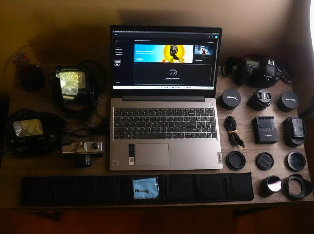 A variety of photography equipment is set on a desk and shot from above. A laptop is open to Photoshop's main page in the middle of the image. On the right side of the image, the desk holds a Canon 5D Mark III, two Ultimax lenses, a Canon 50mm f/1.8 lens, two battery chargers and one battery charger adapter, front and rear caps for the lens being used to take the image, a front cover for the camera being used to take the image, a lens hood, and a stack of graded color filters for the various lenses. Beneath the laptop towards the bottom of the desk is a protective slip case for more color-graded lens filters and a microfiber cloth still in its bag. To the left of the laptop are a Maxim MF-IX film camera, two smart multicolor LED floodlights, and Tone Madison's 2022 MAMA Award Trophy. The desk is wooden and the entire image is bathed in a soft brown light. Just off to the right side of the disk, a sliver of a Neewer ring light is shown illuminating the desk and all of the camera equipment.