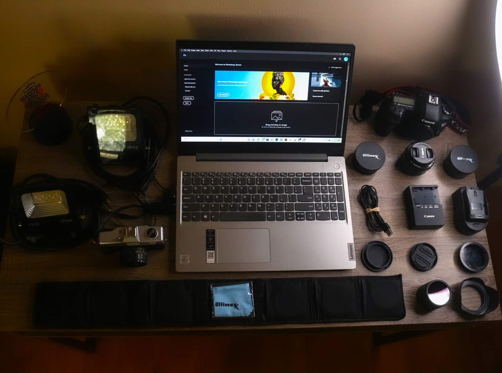 A variety of photography equipment is set on a desk and shot from above. A laptop is open to Photoshop's main page in the middle of the image. On the right side of the image, the desk holds a Canon 5D Mark III, two Ultimax lenses, a Canon 50mm f/1.8 lens, two battery chargers and one battery charger adapter, front and rear caps for the lens being used to take the image, a front cover for the camera being used to take the image, a lens hood, and a stack of graded color filters for the various lenses. Beneath the laptop towards the bottom of the desk is a protective slip case for more color-graded lens filters and a microfiber cloth still in its bag. To the left of the laptop are a Maxim MF-IX film camera, two smart multicolor LED floodlights, and Tone Madison's 2022 MAMA Award Trophy. The desk is wooden and the entire image is bathed in a soft brown light. Just off to the right side of the disk, a sliver of a Neewer ring light is shown illuminating the desk and all of the camera equipment.
