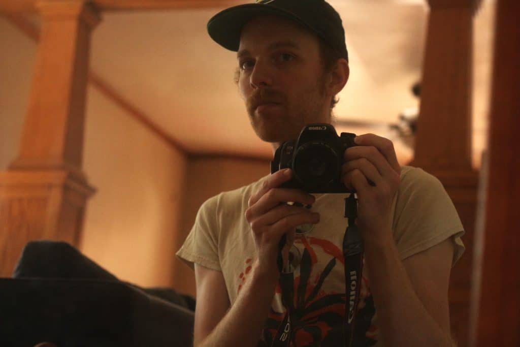 Steven Spoerl is shown holding his Canon Rebel t5i in a white band shirt and green baseball cap. A Canon strap dangles from the lens, which is being held with both hands. He is looking directly into the mirror, and his full face is visible, as the camera only blocks out his neck.