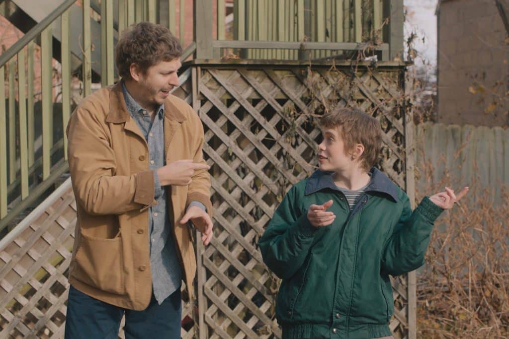 In the backyard of a house, a man in his late twenties wears a tan jacket, looking and pointing at his shorter, younger sister. She wears a green jacket and returns his gaze while making an exaggerated gesture with her hands.