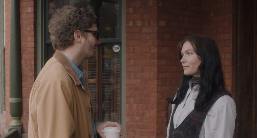 A man in his late twenties wears sunglasses and a tan jacket. He stands on a porch, staring at a woman, also in her twenties, with dyed black hair. She wears a light blue dress shirt and patterned vest, returning his gaze.