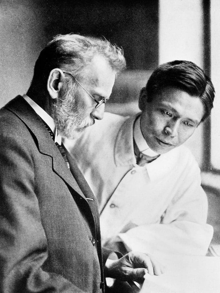 Archival black-and-white photo of an older white man and a younger Japanese man. 