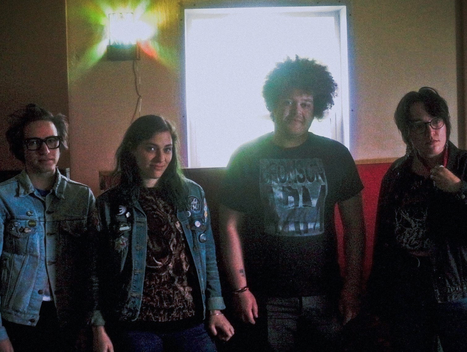 The members of Cult Of Lip pose in Mobius Glen, their studio and practice space. From Left to Right: Ronnie Lee, Hannah Porter, Terrance Barrett, and Emili Earhart. All four members are leaning against a wall, and smiling. Behind them is a window that reveals a wooded area. A stained-glass lamp is lit in the upper right corner. The photo is grainy.
