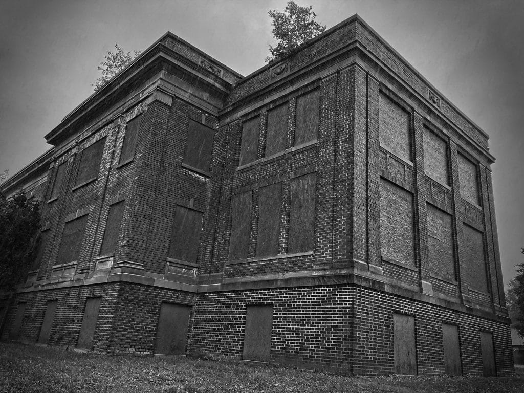 Low-angled black-and-white photo of a brick building with boarded-up windows.