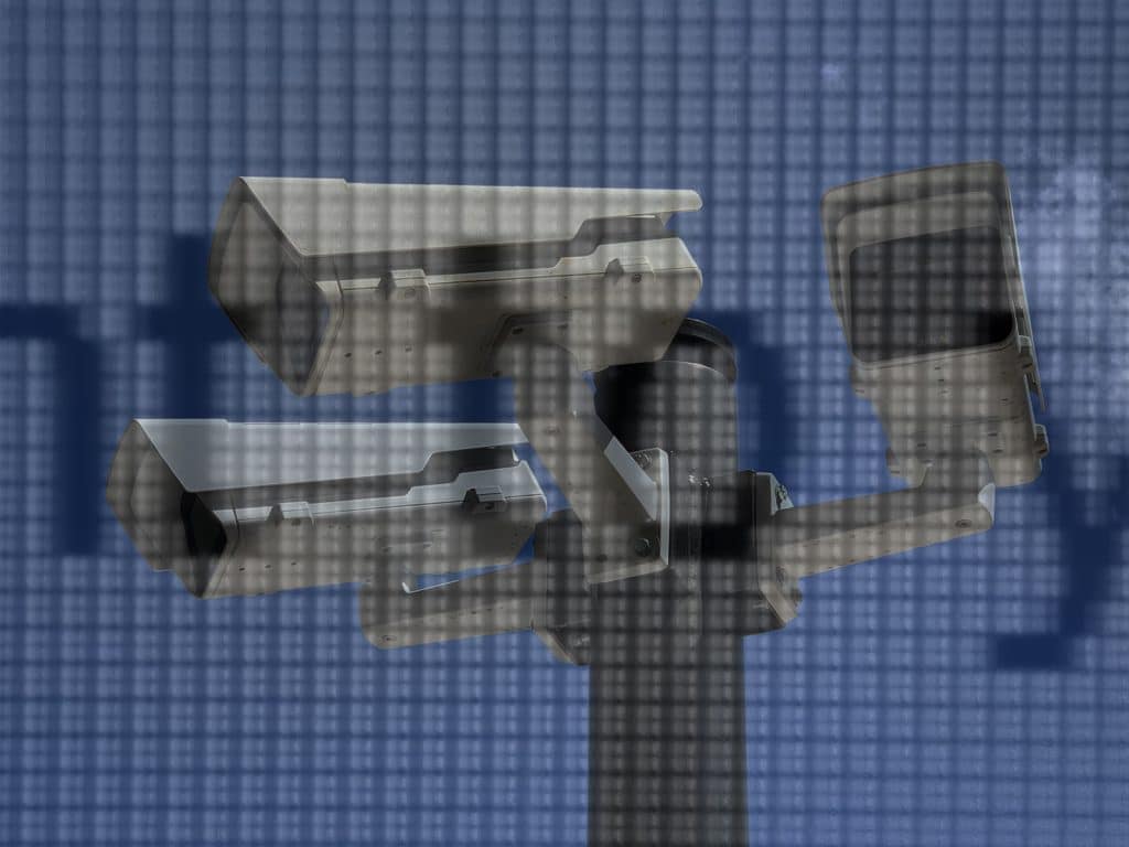 A photo illustration shows a view of three security cameras mounted on a pole with a bright blue sky in the background. Overlaid is a translucent close-up of text on a CRT monitor.