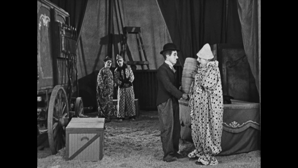 Charlie Chaplin's Tramp character stands next to a clown in the foreground, who is handing him a stack of breakable ceramic plates. Two other clowns stare on from backstage, looking slightly concerned.