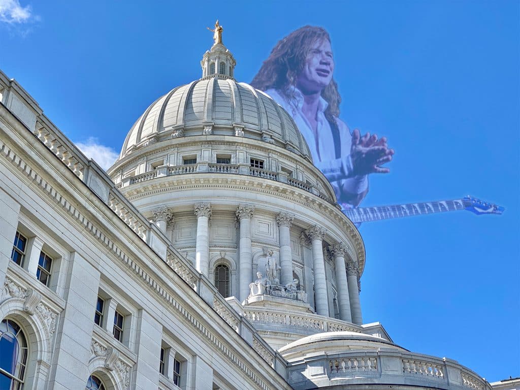 A photo illustration shows a shot of the Wisconsin Capitol dome from below. Behind it, in the sky, is an image of Dave Mustaine wearing a guitar and clapping.