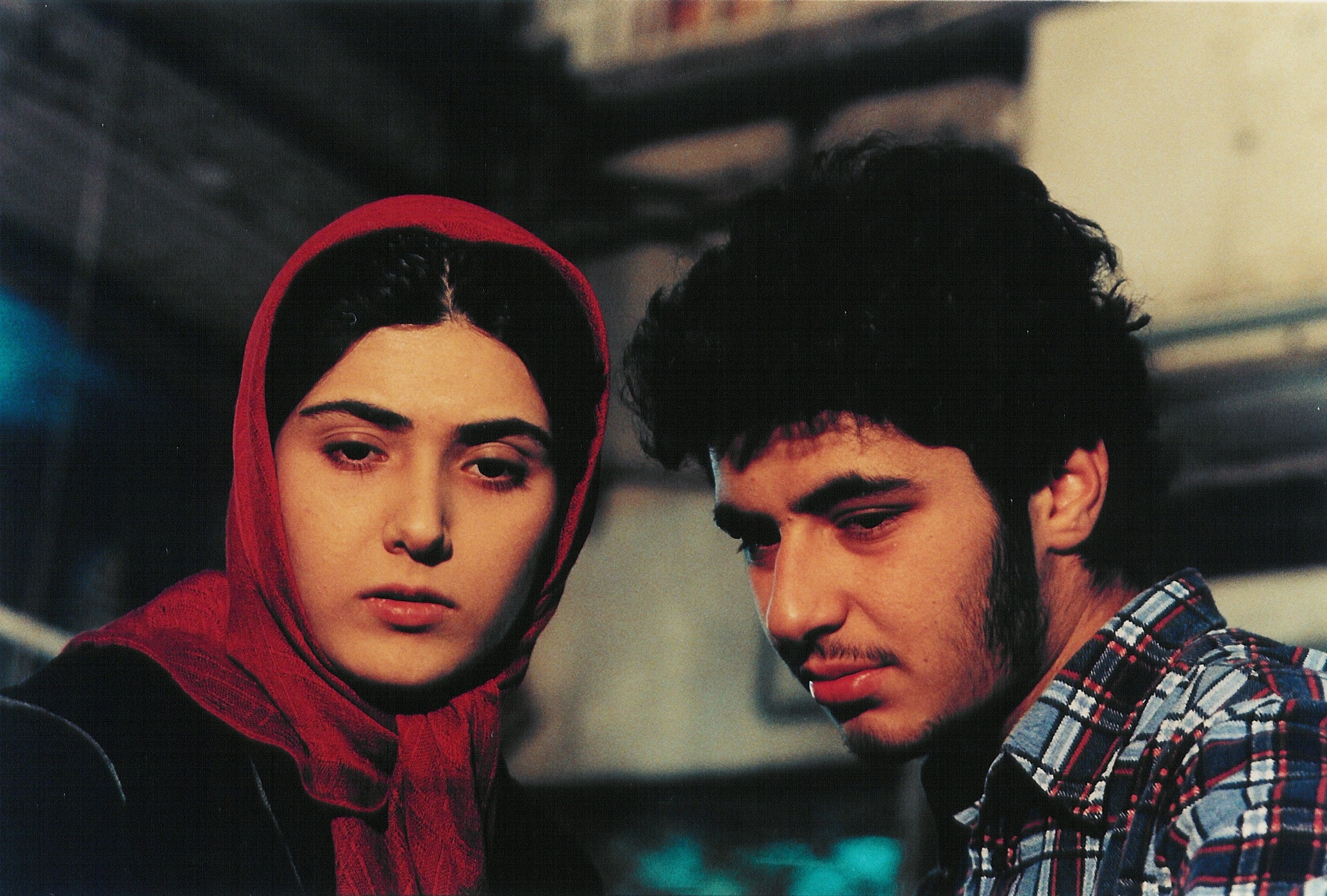 A young Iranian couple with dark hair wearing ambiguous expressions are framed at close-up, and look down at the camera.