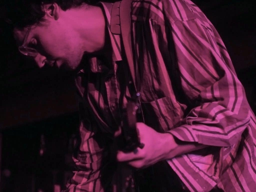 Graham Hunt looks down to the floor as he adjusts a guitar pedal that's out of frame. He's wearing a striped button-up that's unbuttoned at the sleeve and holding a guitar as he kneels down to make the pedal adjustment. The photo is faded, with a twinge of dusty pink.