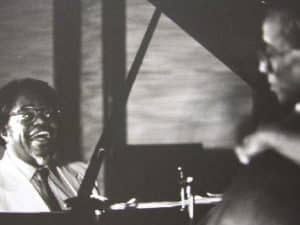 Sir Roland Hanna smiles wide while playing piano. Hanna's looking to his right, at bassist Richard Davis. The two are perforrming together in an old, faded, and slightly blurry black-and-white photograph.