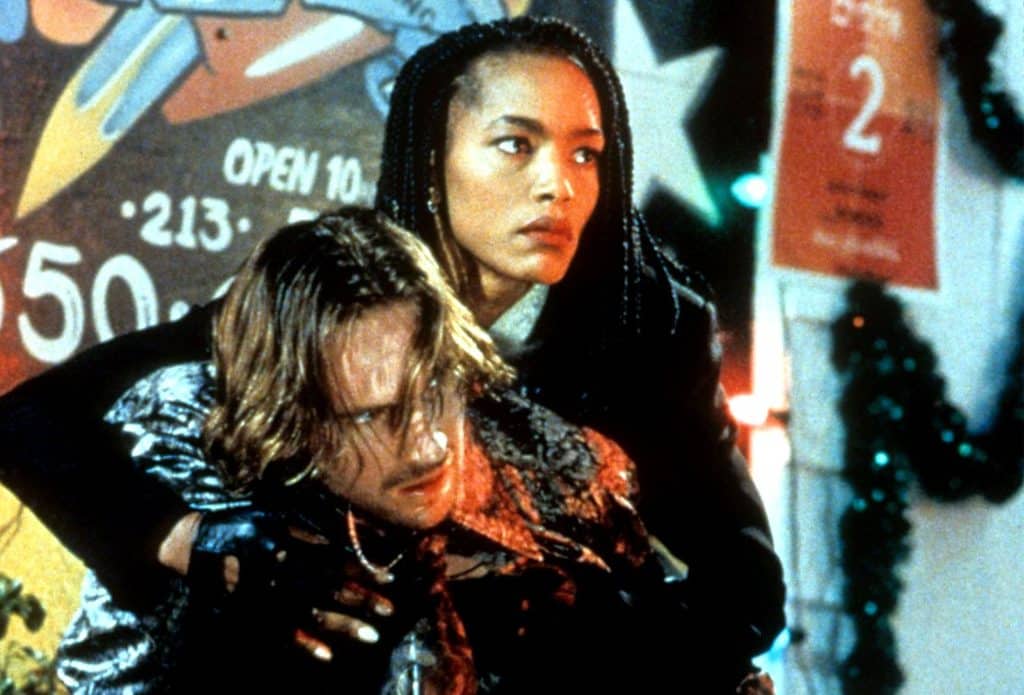 A promotional still from the movie "Strange Days" shows a woman with braided black hair clutching a weakened man with shaggy brown hair.