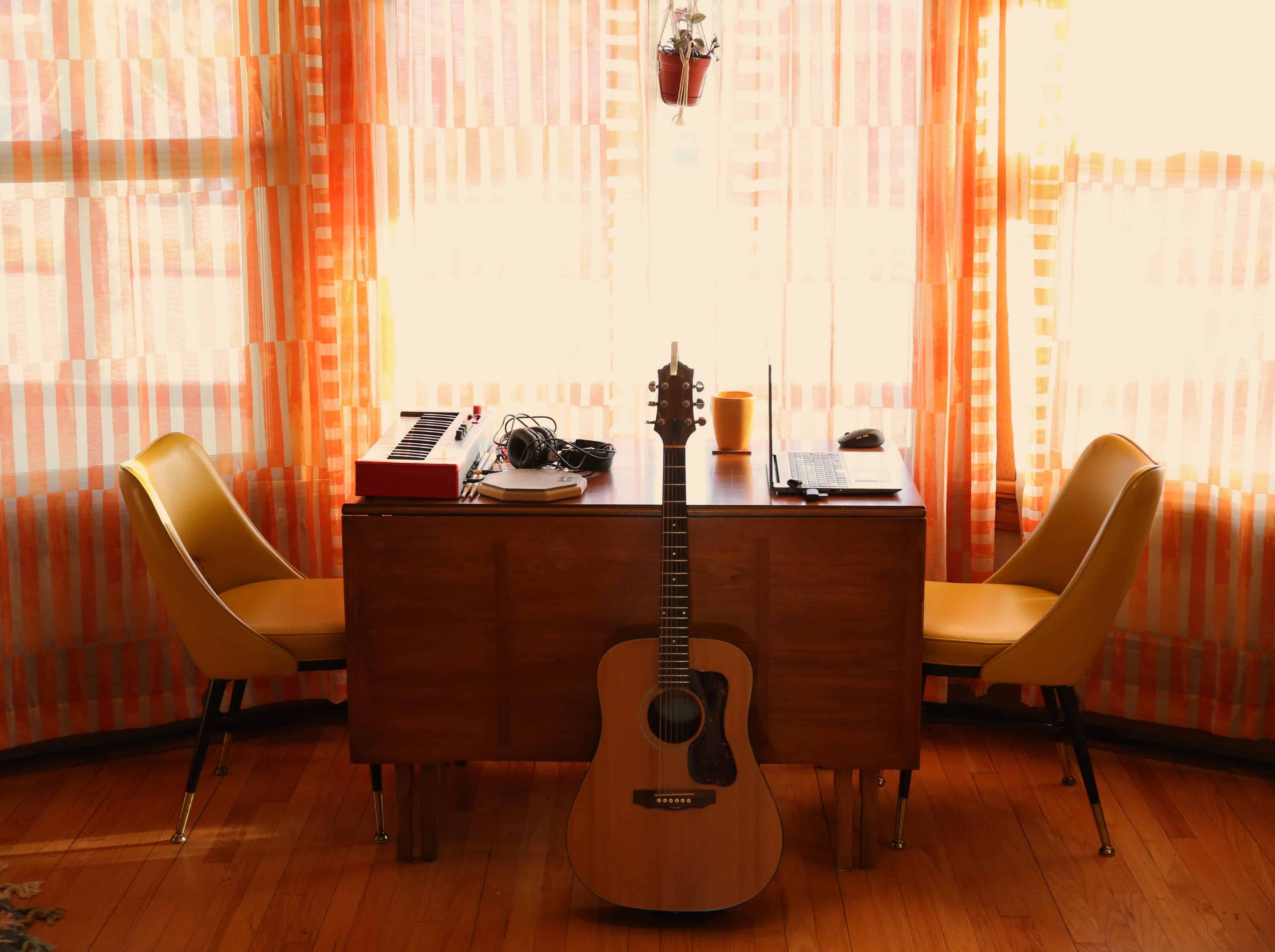 A plant hangs above a table. An acoustic guitar rests against the table, which is centered. Two yellow chairs are on either side. On the right of the table is a laptop, gold coffee mug on a coaster, and a mouse. On the left end is a synth, drum pad, a pair of drumsticks, and studio headphones. Behind the table are large windows covered in semi-transparent drapes that have a light orange and creme vertical bar pattern. The floor is natural wood.