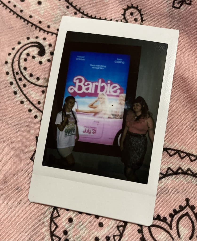 Hanna (right) and Tara (left) stand in front of the "Barbie" poster outside a movie theater. The photo is captured on a slender instant photograph, which lays on top of patterned salmon-colored fabric.