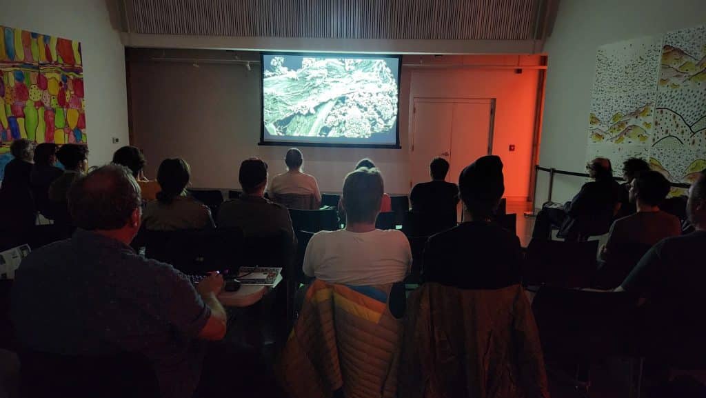 A audience of approximately 20 sits in a darkened room that also doubles as an art gallery to watch a short film digitally projected on a screen.