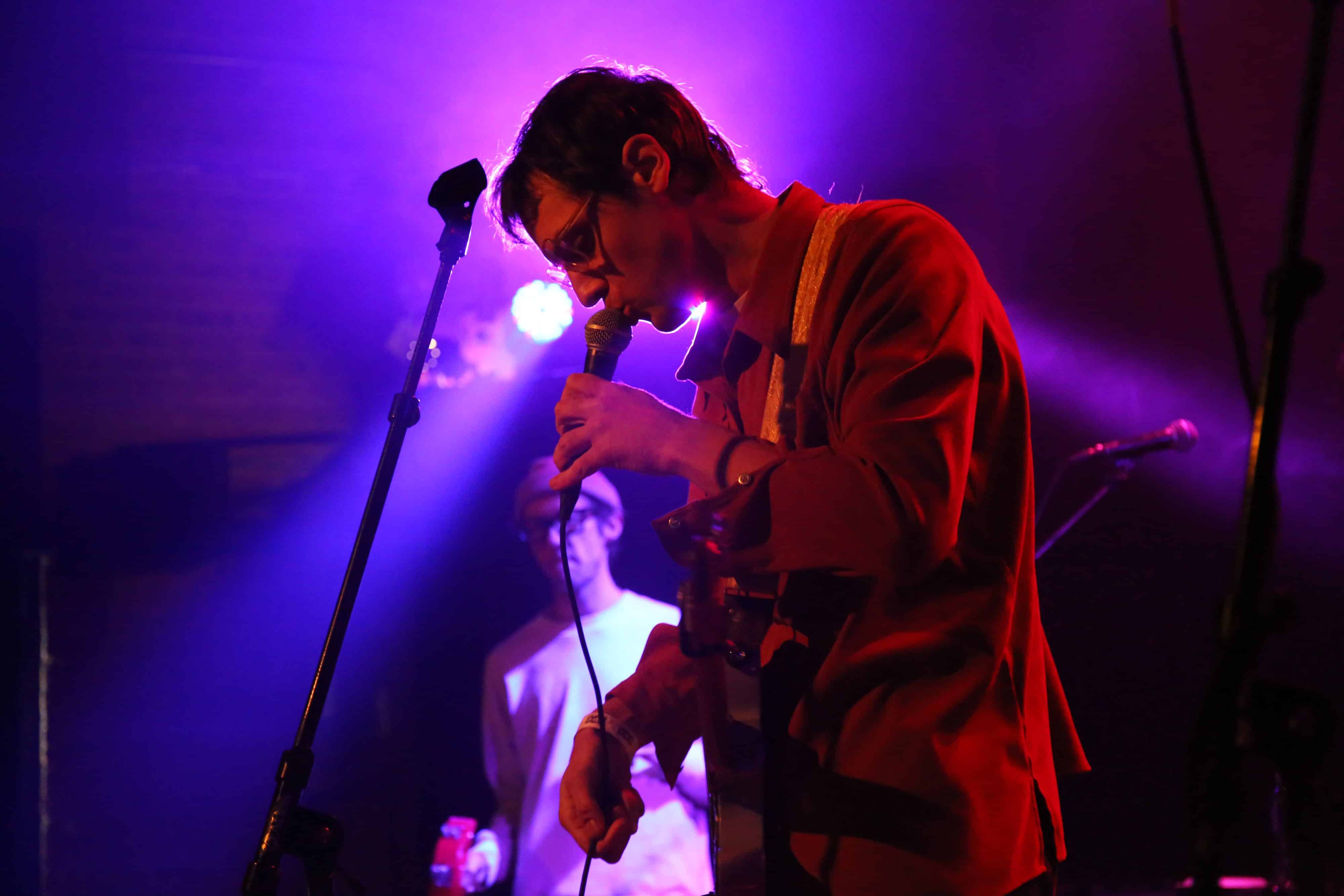 Graham Hunt performs live at the High Noon Saloon. He's wearing a red button-up long sleeve shirt and gripping a microphone with his left hand and singing with his head down. His right hand grips the mic cable. Behind him is a cascade of blue and purple stage lighting. The stage lighting provides a soft, halo illumination effect that surrounds Hunt's head. Dusk bassist Ridley Tankersley can be seen in soft focus several yards behind Hunt, playing tambourine.