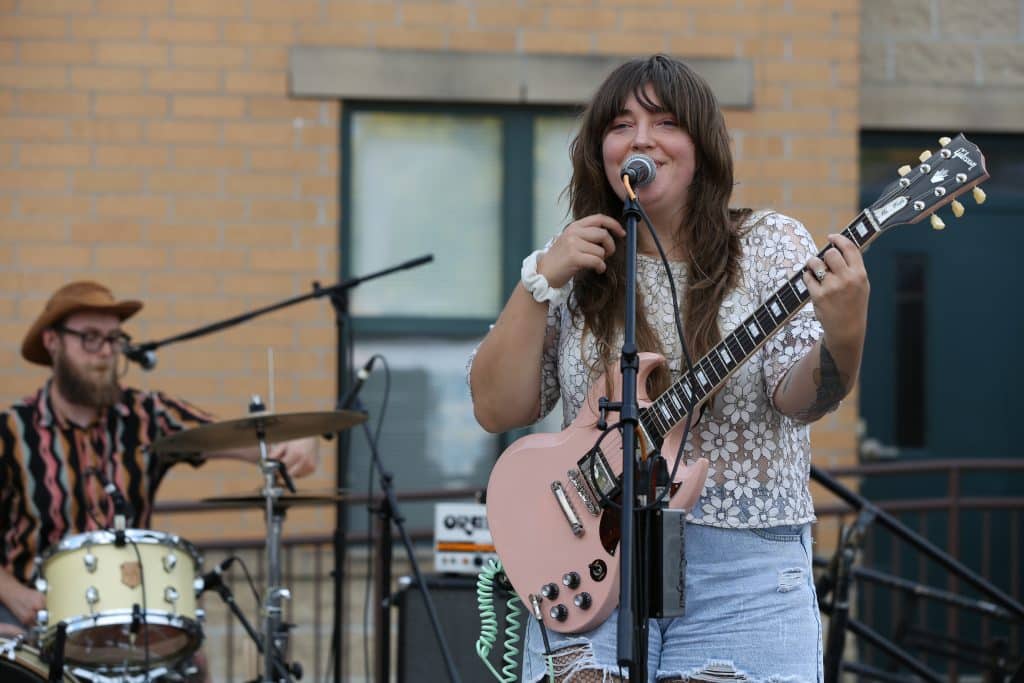 Lunar Moth perform on the WORT stage at the Willy Street Fair. There's a pale-brick building in the background. To the right of the image, guitarist/vocalist Amber Fasula is playing guitar and smiling while singing into a microphone. She's wearing a sheer white lace flower print shirt and a frayed denim skirt. Behind her to the left of the image, Mac Felckowski sets up a microphone at his drumset. He's wearing a striped button-up shirt and a cowboy hat.