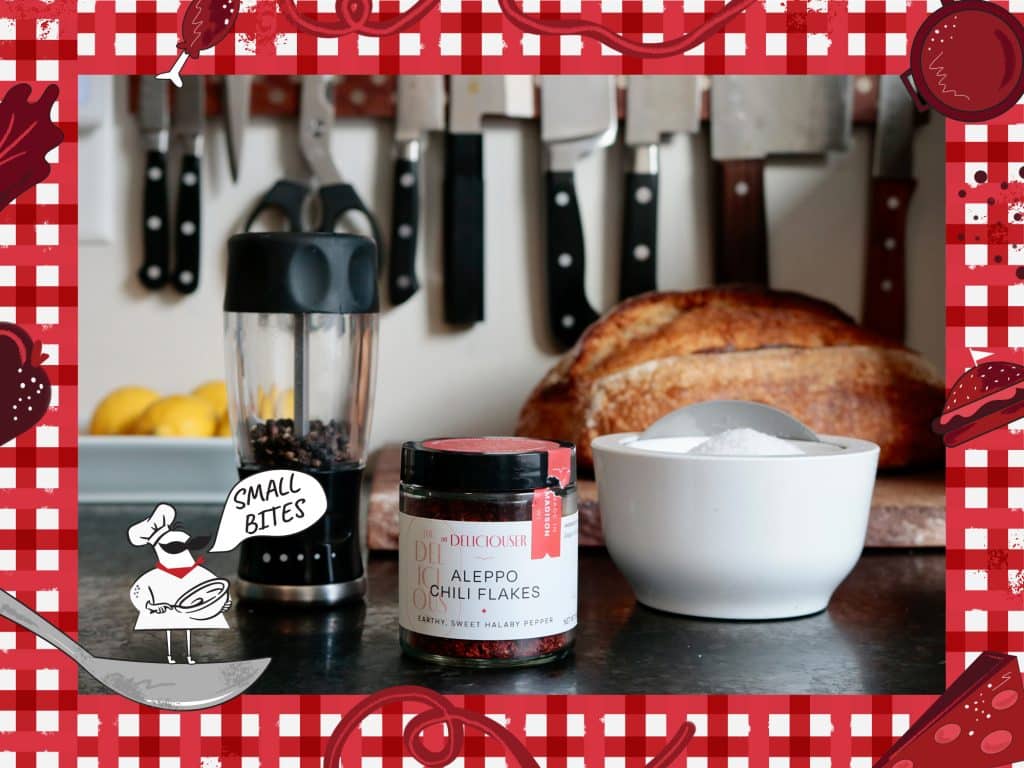 A photo shows an Aleppo chili flakes jar flanked by salt and pepper on a kitchen counter. An illustrated frame around the photo depicts a checked tablecloth pattern with various items of food. In the bottom left corner, a small caricature chef stands on a spoon with the speech bubble, "Small Bites."