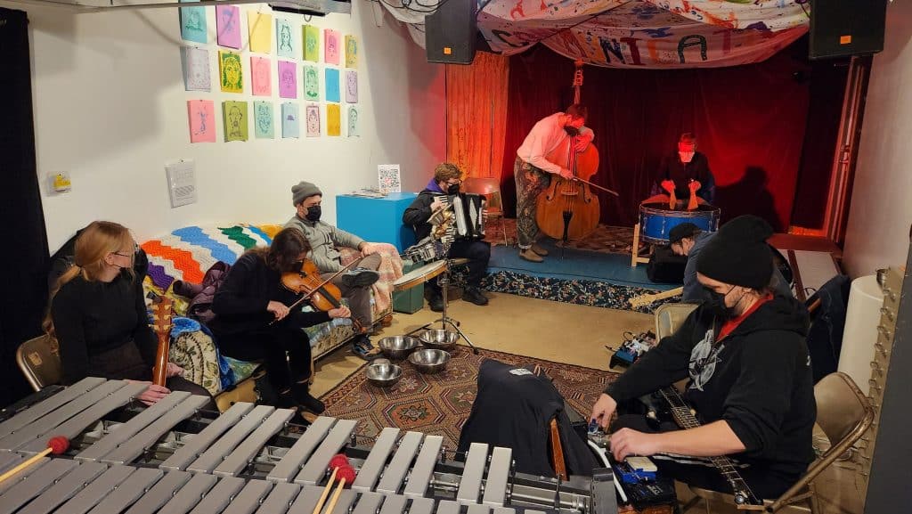 Several musical performers sit in a rough rectangular shape facing each other during a jam session at Communication, an all-ages arts gallery and performance space. The stage at the back of the room is mood-lit in red, while the rest of the room is more brightly lit in white fluorescents.