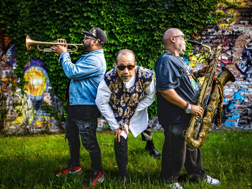 A photo shows the three current members of the Ethnic Heritage Ensemble posing against a wall covered with ivy and colorful paintings. In the center is Kahil El'Zabar, crouched and facing the camera. To either side of him are trumpeter Corey Wilkes (left) and saxophonist Alex Harding (right). Wilkes and Harding both stand with their backs facing El'Zabar, with their respective instruments lifted up to their lips.