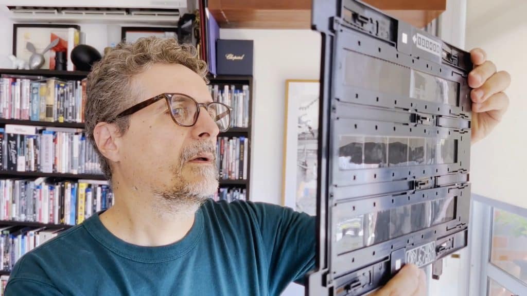 A man in his late 40s, wearing glasses, holds up strips of photographic negatives to the sunlight to look more closely. He stands in a room with several shelves of DVDs and Blu-rays behind him out of focus.