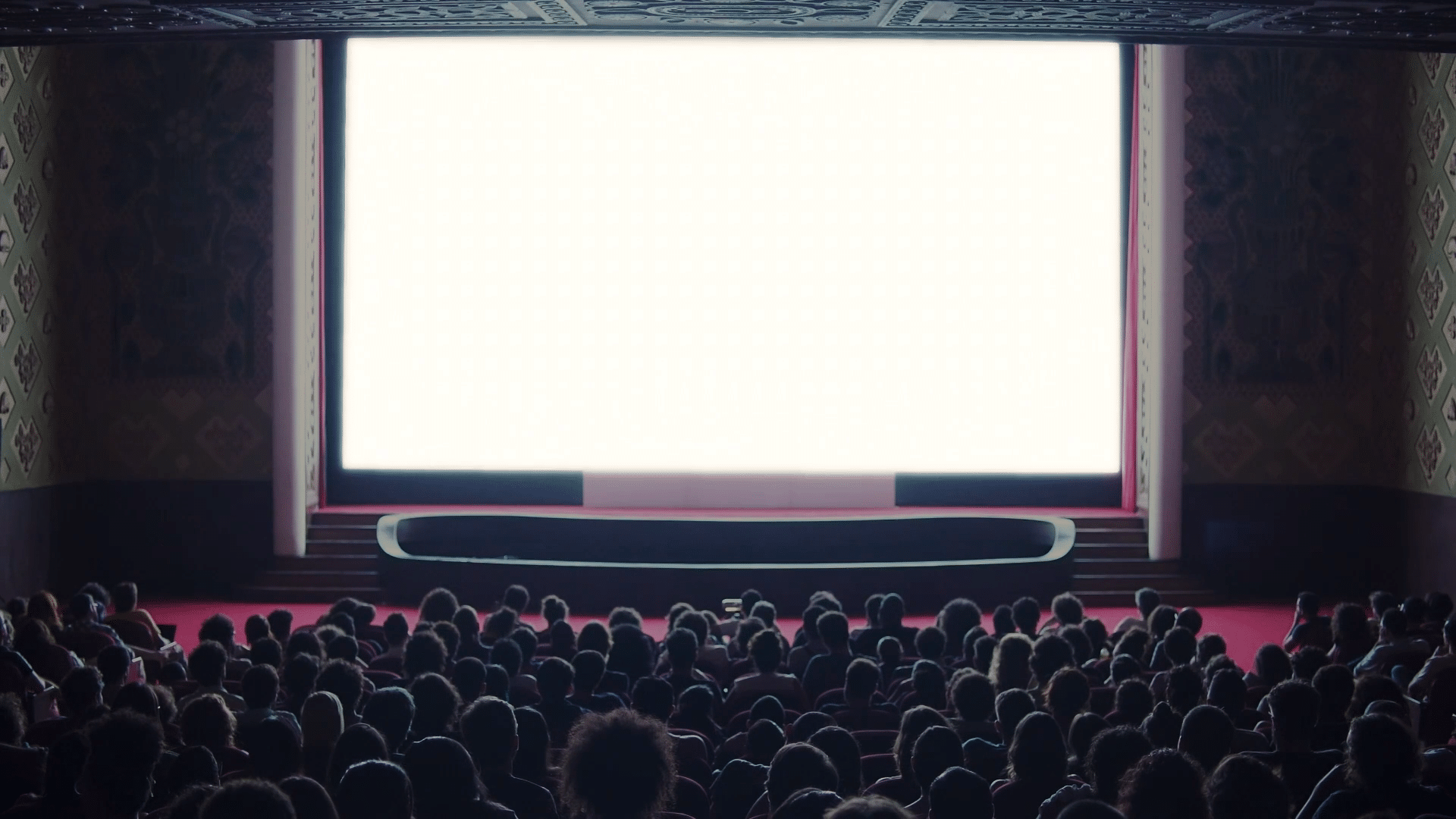 A packed audience in the foreground faces towards a large movie screen in front of them that is glowing bright white.