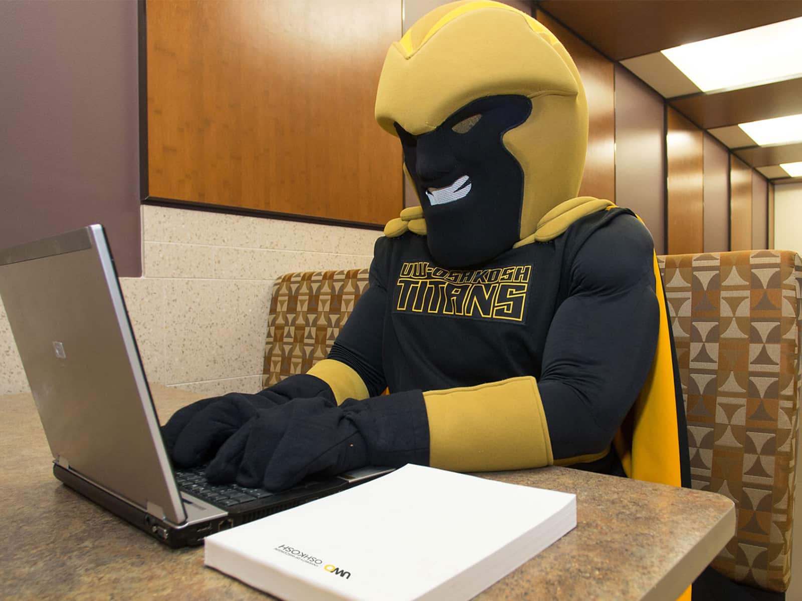 A photo shows UW-Oshkosh’s mascot, Clash, sitting in a booth in a campus building, working on a laptop computer. Clash is a figure dressed in black with a gold cape, helmet, and cuffs, and a shirt that reads “UW-Oshkosh Titans.”