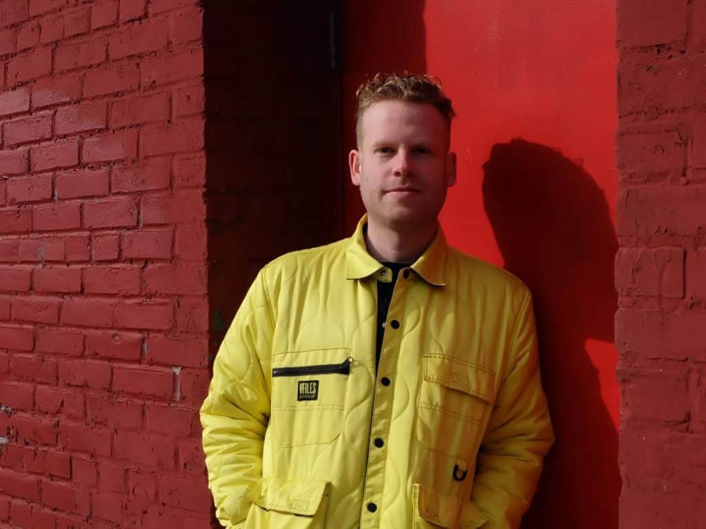 Jonah Parzen-Johnson is pictured leaning against a red door tucked into a red brick building. He's just right of center in the image, wearing a bright yellow jacket. His face is partially covered in shadow, with the shadow of his body being cast onto the door behind him. He is smiling. 