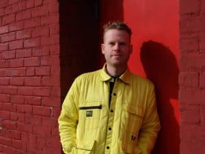 Jonah Parzen-Johnson is pictured leaning against a red door tucked into a red brick building. He's just right of center in the image, wearing a bright yellow jacket. His face is partially covered in shadow, with the shadow of his body being cast onto the door behind him. He is smiling.