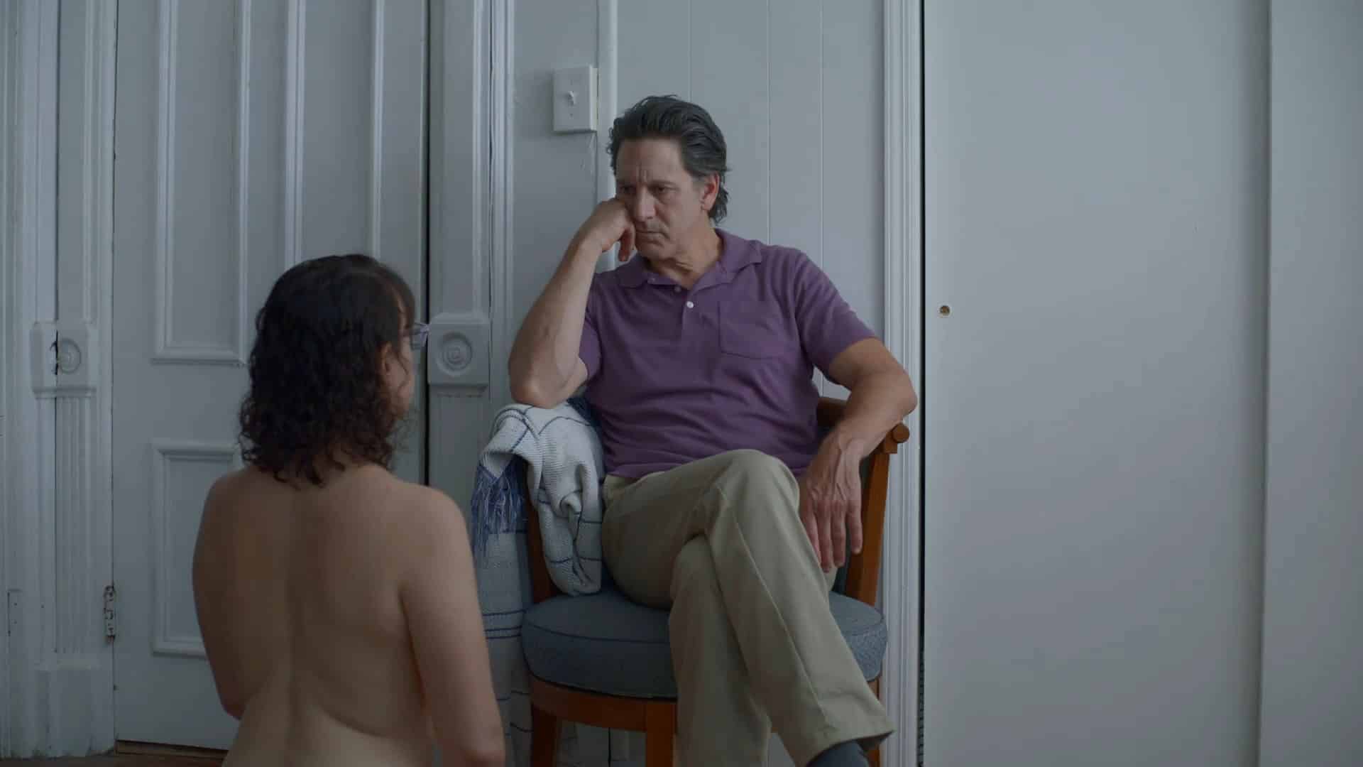 A thirtysomething woman (Joanna Arnow) with mid-length long hair kneels submissively (and shirtless) on the floor in a white-walled bedroom. She faces away from the camera and towards her partner, an older man (Scott Cohen), who is fully dressed, sitting in a chair and staring intently at her.