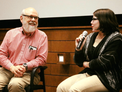 In front of a movie theater screen and wooden panels below it, two people sit and look at each other in conversation. David Bordwell, left, wears a red and white plaid shirt, turning towards Kris Johnson-Salazar in a black and white cardigan, who holds the microphone. Both smile.