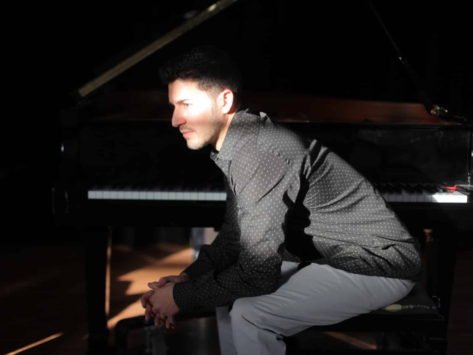Emmet Cohen is shown in profile on a piano bench, hunched over and clutching his hands together. A piano is visible just behind him. The image is dark but a spot of light illuminates his face, creating a contemplative mood. He's wearing light blue jeans and a black button-up long-sleeve shirt with a pattern of small white markings.