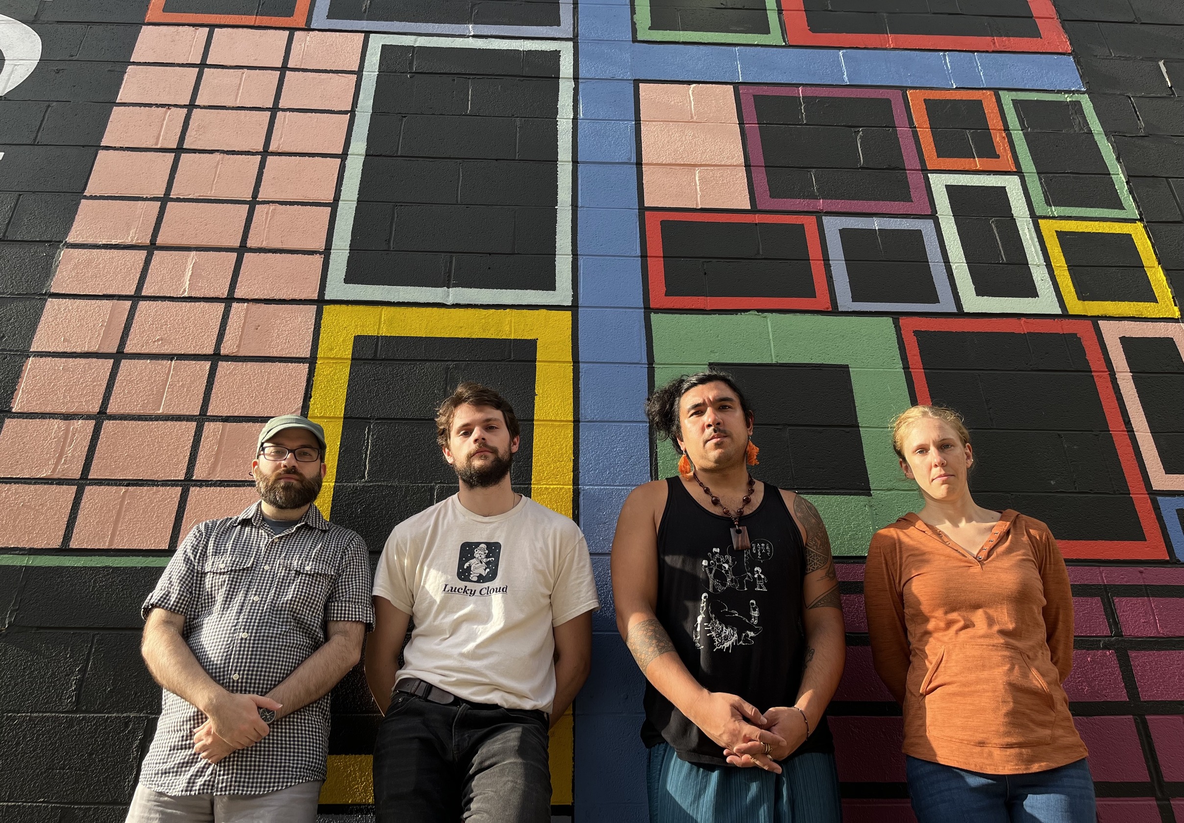 The four members of the Jakob Heinemann Quartet lean against a wall painted with rectangles in numerous sizes and colors, both shaded and outlined. The members all face the camera with neutral expressions, as the lens distorts at the edges of this sunny, low- and wide-angled shot.