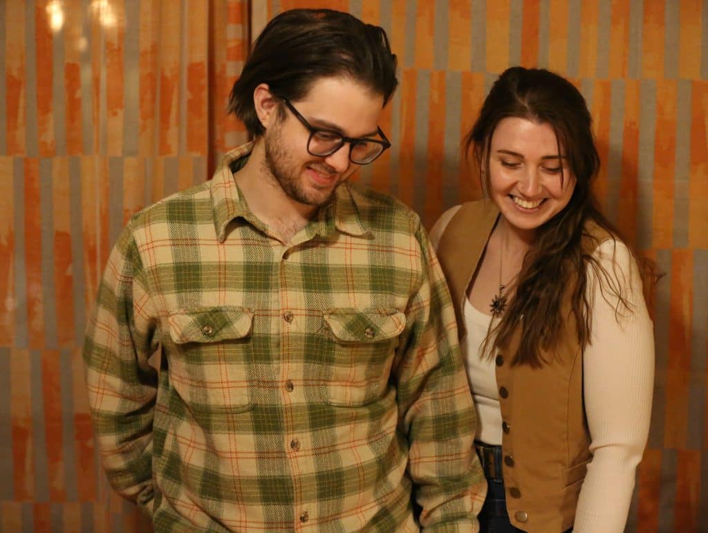 Marley Van Raalte (Left) and Abby Self (Right) are standing together, looking down out of frame and smiling. An orange-cream drape curtain is behind them. Van Raalte is in a pea-green plaid with small red accents and Self's wearing a brown vest, necklace, and white long-sleeve shirt.