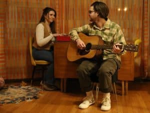 Abby Self (Left) and Marley Van Raalte (Left) are looking directly at each other as they play music around a living room table. Van Raalte is looking over his shoulder and Self is smiling.