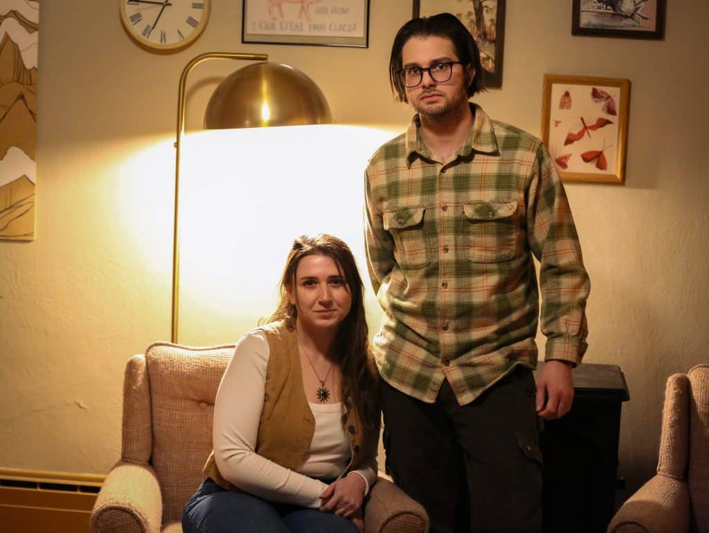 Abby Self (Left, seated) and Marley Van Raalte (Right, standing) both look directly at the camera. A directional lamp casts a halo light effect around Self. Around Van Raalte's head-height are a series of eccentric art prints and a clock.