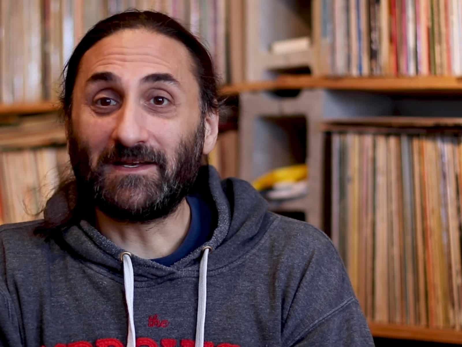 Aadam Jacobs, the subject of the film "Melomaniac," smiles excitedly in a still from the film. He is to the left of the image in a gray hoodie with white drawstrings. Visible behind him are two fully stocked record shelves, running along two walls. Jacobs' long black hair is pulled back, and he sports a full beard.