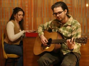 Loveblaster's Abby Self (Left) and Marley Van Raalte (Right) play music together in a living room. They are both seated. Self is in soft focus, playing synth, while Van Raalte is closer to the camera, playing acoustic guitar. Both musicians are seated around a table. Behind them is an orange-cream drape.