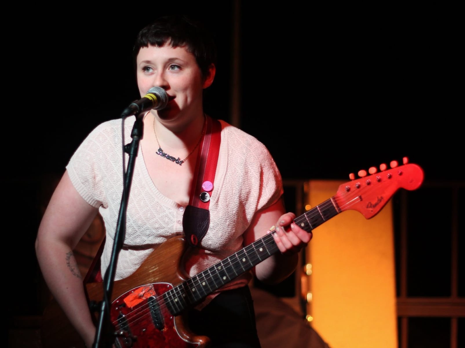 Allison Crutchfield of the band Swearin' playing guitar and singing at UW's Memorial Union Terrace in May 2014. She's wearing dark pants and a white shirt with a low neckline. A sparkly necklace that says "Swearin" is visible. Her hair is dark and in a pixie cut. She's smiling.