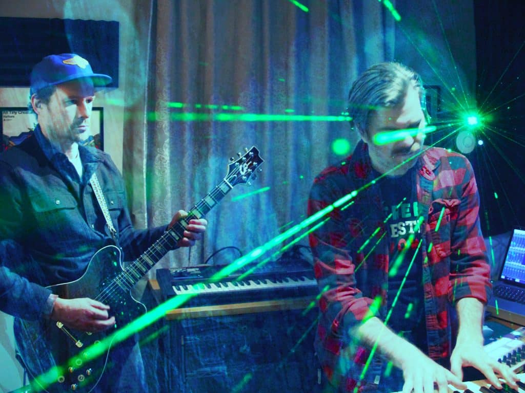 A photo shows All Tiny Creatures members Andrew Fitzpatrick (left) and Thomas Wincek performing together in an indoor space. Fitzpatrick is standing and playing a guitar, and Wincek is standing and playing a keyboard. Other devices, including keyboards and a laptop, are arranged around them. Streaks of green light radiate across the photo.
