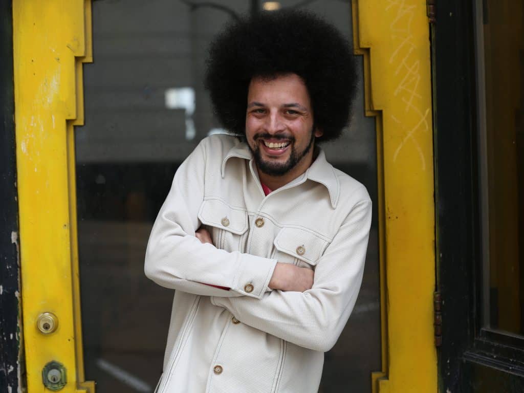 Kevin Willmott II is wearing a white button-up coat. He's smiling, sporting a well-maintained beard and afro. His arms are crossed. He's posing in front of the front door of Gamma Ray Bar, his new business.