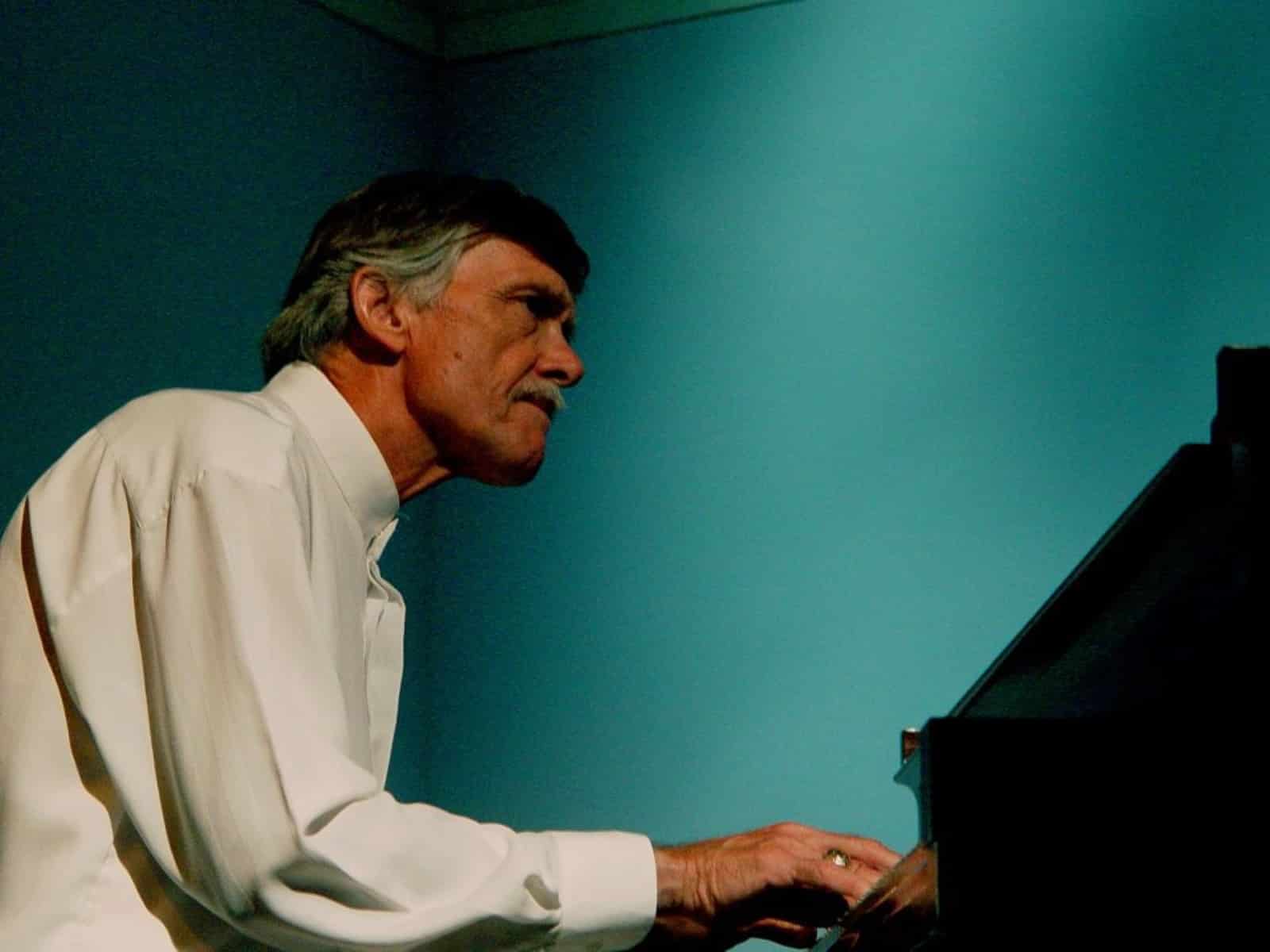 John Harmon is shown playing piano. His expression conveys a sense of deep focus. He's wearing a white button-up dress shirt and a wedding ring is visible on his ring finger as he plays. The image seems to be shot from below, making Harmon look larger than life. His mustache and hair are gray. The top of his head is obscured by shadow. The photo is grainy and there is a light blue-green hue to the image as a result of the overhead stage lights.