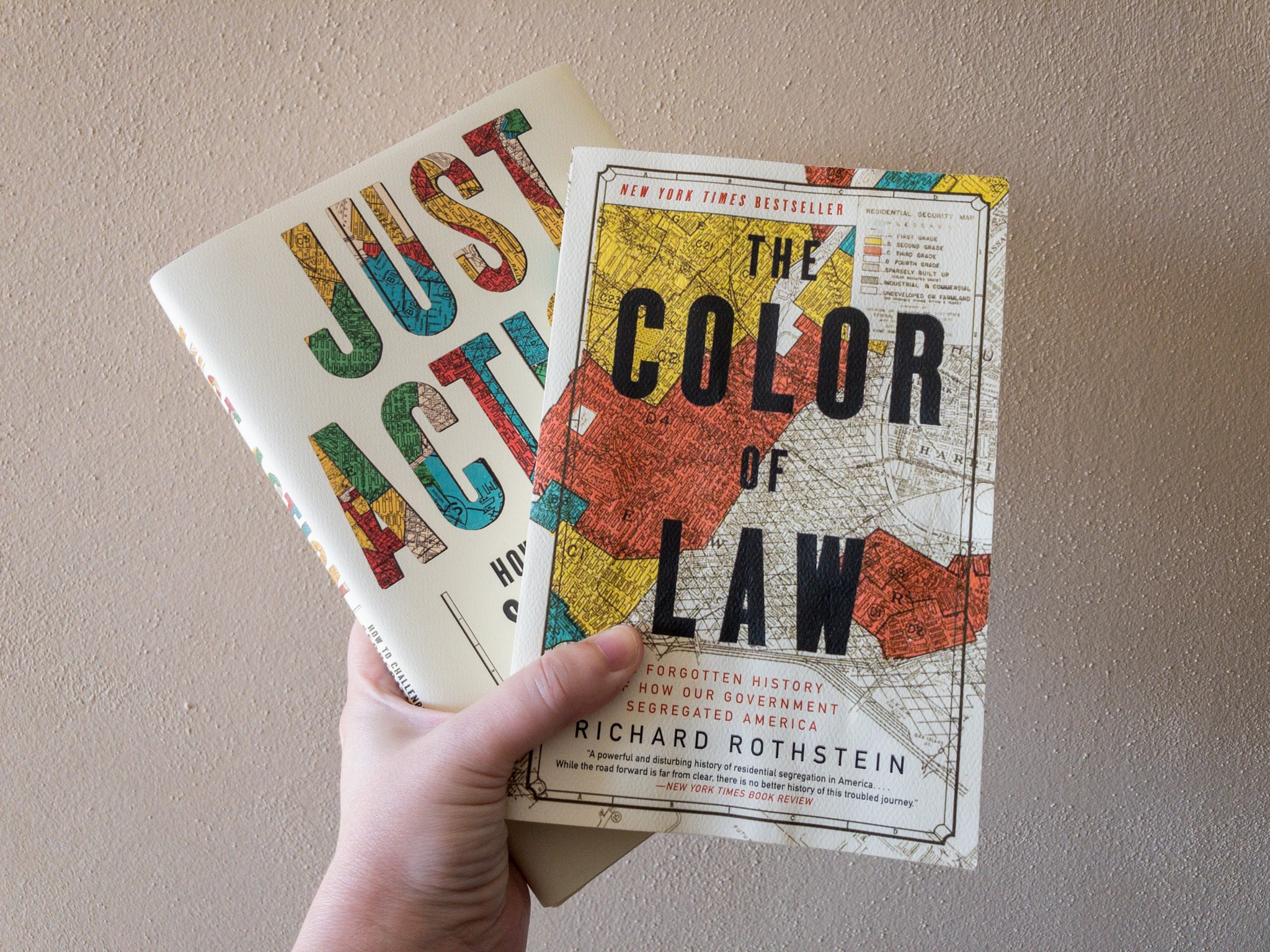 Photo of the author’s hand holding paper and hardback copies of "The Color of Law" and "Just Action" in front of a beige wall.