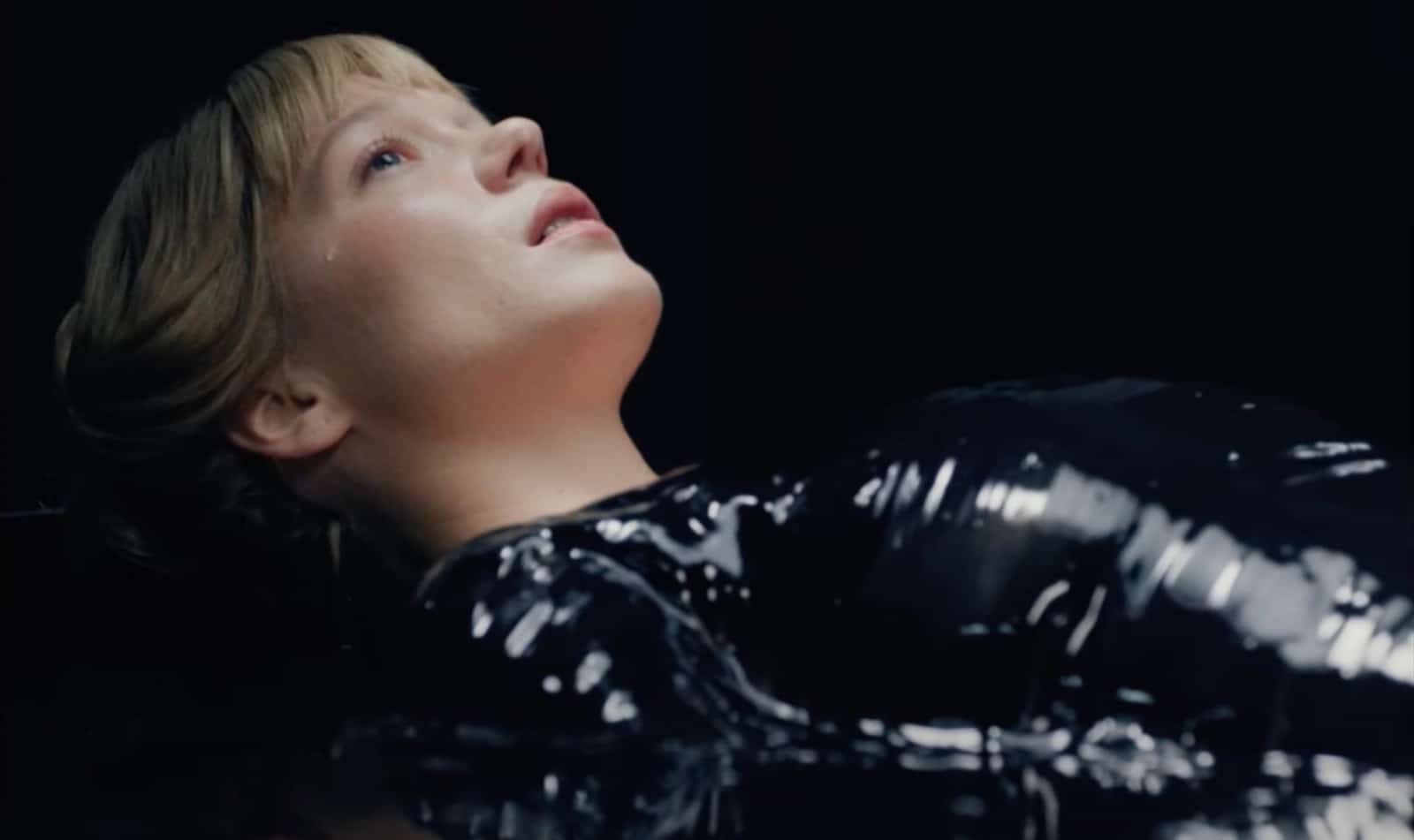 A blonde woman lays face-up in partial profile. She sheds a tear, and stares upward in a black, blank space. Her body is wrapped in a similarly black-colored substance.