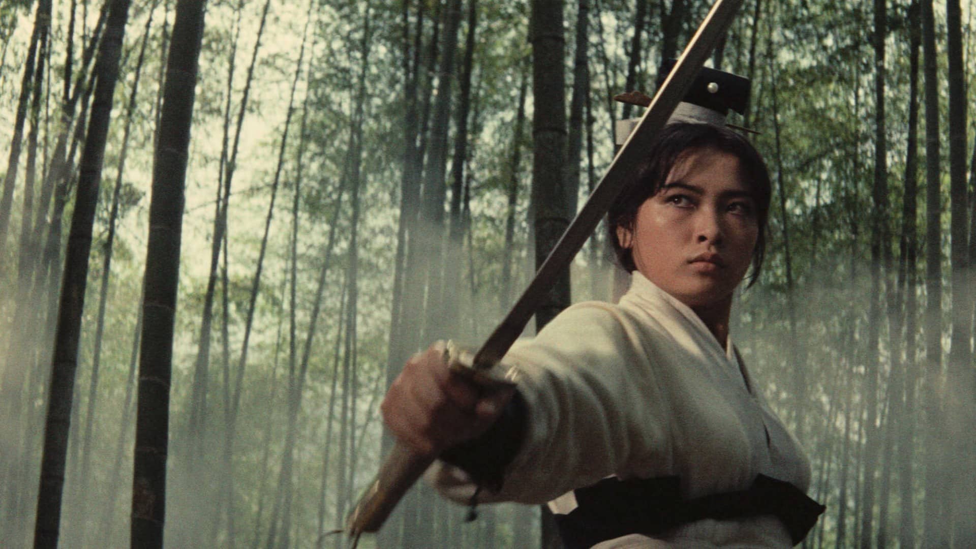 A woman in a white kimono brandishes a sword. She stands, posed at an angle, with a determined look on her face amid a foggy forest of bamboo reeds behind her.