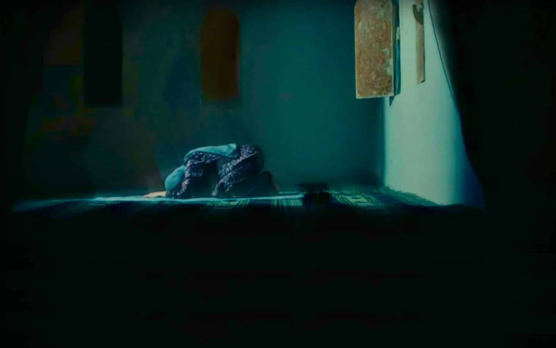 An impressionistic image of a woman kneeling in prayer on her prayer mat is shown in the upper half of a still frame from Def Sonic's music video for "Zephyr." An extensive black border outlines the majority of the image, while the central figure is shown in pale greens and blues, in full dress, and accentuated by artificial lens flaring.